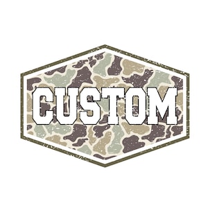 Op de afbeelding: Een verweerd, zeshoekig embleem met het woord "CUSTOM" in vette, witte blokletters met een zwarte omlijning. De achtergrond heeft een camouflagepatroon in tinten bruin, groen en beige.