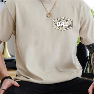 May include: Beige t-shirt with a camouflage "DAD" patch on the chest. The patch is a hexagon shape with a white border. A gold necklace with a circular pendant is visible.
