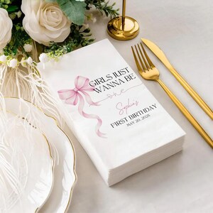 May include: White paper napkins with a pink bow and the text "GIRLS JUST WANNA BE one" and "Sophia's FIRST BIRTHDAY MAY 20, 202X". Gold fork and knife are on the right. White roses and a gold candle holder are in the background.