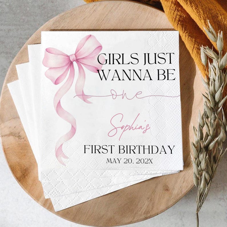 May include: White paper napkins with a pink bow and the text "GIRLS JUST WANNA BE one Sophia's FIRST BIRTHDAY MAY 20, 202X". The napkins are stacked on a wooden tray.