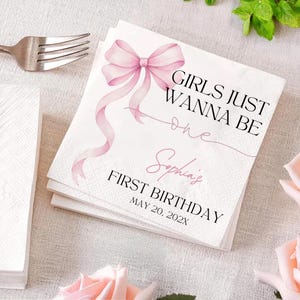 May include: White paper napkins with a pink bow and the text "GIRLS JUST WANNA BE one Sophia's FIRST BIRTHDAY MAY 20, 202X". A silver fork is visible in the upper left corner. Pink flowers are in the lower right corner.