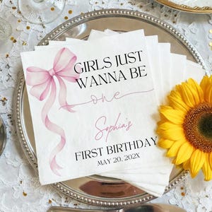 May include: White paper napkins with a pink bow and the text "GIRLS JUST WANNA BE one Sophia's FIRST BIRTHDAY MAY 20, 202X". A yellow sunflower is on the right side of the napkins, which are on a silver plate.