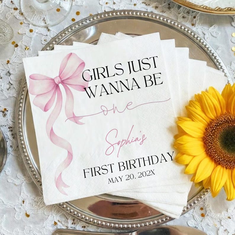 May include: White paper napkins with a pink bow and the text "GIRLS JUST WANNA BE one Sophia's FIRST BIRTHDAY MAY 20, 202X". A yellow sunflower is on the right side of the napkins, which are on a silver plate.