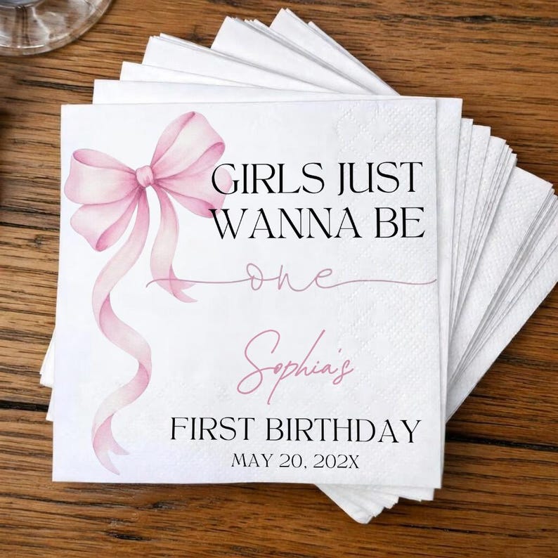 May include: White paper napkins with a pink bow and the text "GIRLS JUST WANNA BE one Sophia's FIRST BIRTHDAY MAY 20, 202X". The napkins are stacked on a wooden surface.