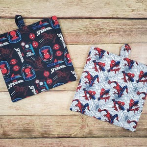 May include: Two square pot holders with loop handles. One is black with red, blue, and white Spider-Man graphics and text. The other is light gray with a red Spider-Man pattern and a web design.