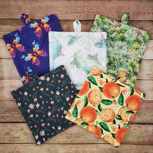 May include: Five square quilted pot holders featuring distinct patterns: a dark blue butterfly print, a white hummingbird design, a green succulent motif, a dark gray floral pattern, and a yellow fabric with a vibrant orange fruit print.