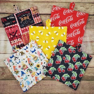 May include: Five square quilted pot holders featuring various patterns: a black and red BBQ theme, a red Coca-Cola logo print, a yellow bee print, a white gnome and sunflower print, and a black fabric with a red cherry and white polka dot pattern.