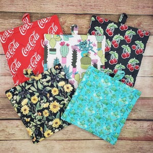 May include: A collection of quilted pot holders with various patterns. Designs include a red Coca-Cola print, cacti, cherries on black, sunflowers, and a turquoise frog pattern. Each pot holder has a loop for hanging.