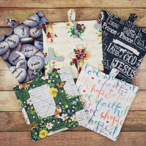 May include: Five square fabric pot holders with hanging loops. One features gray stones with words like Faith and Peace. One has a cream background with crosses and flowers. One is black with white text including Lamb of God. One shows a fox and butterfly. One has colorful text reading faith and hope.