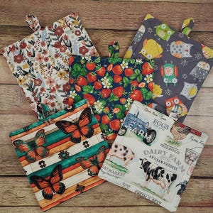 May include: Five square fabric pot holders featuring various prints: a floral pattern, a strawberry design, an owl motif, a butterfly and striped pattern, and a farm scene with a cow, pig, chicken, and text reading Eggs for Sale.