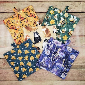 May include: Five square fabric pot holders featuring various prints: one with orange butterflies on yellow, one with bees and flowers on green, one with cats on beige, one with beehives on dark blue, and one with purple wildflowers on a dark blue background.