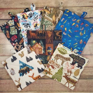 May include: A collection of quilted pot holders with various outdoor-themed patterns. Designs include camping scenes, cabins, fishing lures, and wildlife such as bears, deer, and squirrels. Each pot holder has a loop for hanging.