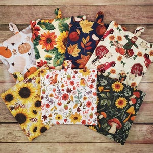 May include: A collection of seven square quilted pot holders featuring various autumn-themed prints, including sunflowers, pumpkins, red barns, farm trucks, mushrooms, acorns, and birds, each with a small fabric loop for hanging on a kitchen hook.