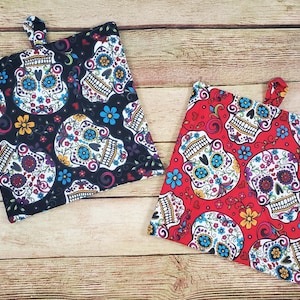 May include: Two square pot holders with a Day of the Dead sugar skull pattern. One is black with white, blue, yellow, and red skulls and flowers. The other is red with the same design. Each has a fabric loop.
