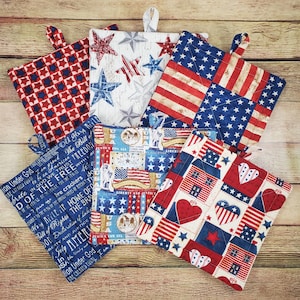 May include: A collection of five square fabric pot holders featuring patriotic themes in red, white, and blue. The designs include star patterns, American flag motifs, text about liberty and freedom, and heart shapes, each equipped with a fabric hanging loop.