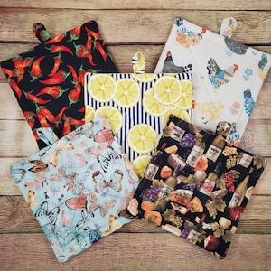 May include: Five square fabric pot holders with hanging loops. Patterns include red chili peppers on black, yellow lemon slices on blue stripes, colorful chickens on white, butterflies on light blue, and wine bottles with grapes on black.