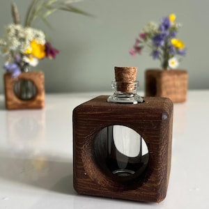 May include: A small glass bottle with a cork stopper sits on a dark brown wooden cube with a circular opening. The cube is part of a set of three, each holding a small bouquet of colourful wildflowers. The background is a soft green.