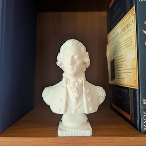 May include: A white bust of the Marquis de Lafayette featuring a detailed 18th-century military uniform with epaulets and a cravat. The name Lafayette is embossed on the front of the base. The sculpture is positioned between two dark blue books.