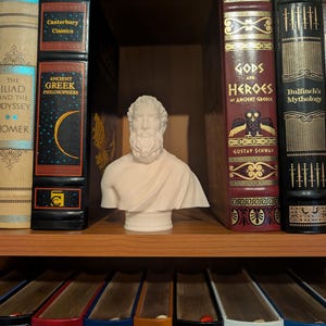 Può includere: Un busto bianco di un uomo barbuto è posto su una libreria, circondato da letteratura classica. I libri hanno lettere dorate e dorsi decorativi. I titoli includono "L'Iliade e l'Odissea", "Antichi filosofi greci" e "Dei ed eroi dell'antica Grecia".