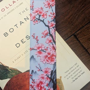 以下が含まれることがあります： ピンク色の桜と灰色の枝が描かれた、明るい青色の背景の長方形のブックマーク。 ブックマークは、「The Botany of Desire」というタイトルの本の上に置かれています。