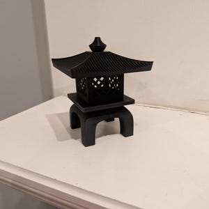 Puede incluir: Una linterna pagoda japonesa negra en miniatura con un techo de varios niveles y lados calados. La linterna se asienta sobre una base cuadrada sostenida por cuatro patas curvas. La pieza decorativa está hecha de un material oscuro y sólido.