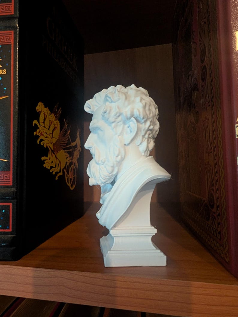 May include: A white bust of a bearded man with curly hair, set on a pedestal. The sculpture is positioned on a wooden shelf between two books with gold lettering and decorative covers. The bust is a classical art piece.