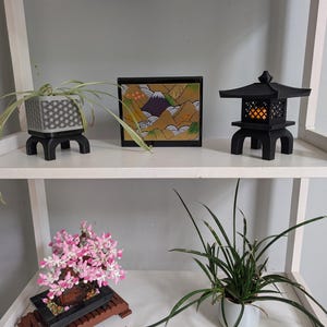 Puede incluir: Una estantería blanca exhibe una decoración de temática japonesa. Los artículos incluyen una linterna negra, una pintura de paisaje enmarcada y una maceta con estampado geométrico con una planta. También hay un bonsái con flores rosas y una planta en maceta blanca con hojas verdes largas.
