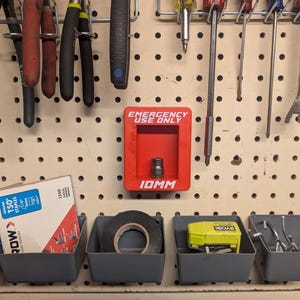 以下が含まれることがあります： 工具と収納容器を備えた木製のペグボード。ペンチ、ドライバー、バールが見えます。「EMERGENCY USE ONLY」と書かれた赤い箱が取り付けられています。グレーのビンには、ホッチキスの針、テープ、電動工具、その他のハードウェアが入っています。