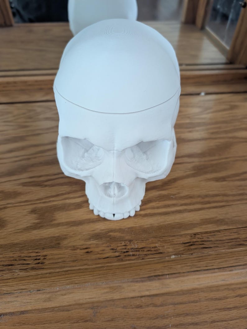 Puede incluir: Un objeto blanco con forma de calavera con rasgos faciales detallados, que incluyen cuencas de los ojos, una cavidad nasal y dientes. La calavera descansa sobre una superficie de madera, con una segunda calavera visible en el fondo.