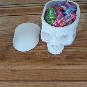 Puede incluir: Un recipiente blanco con forma de calavera con tapa extra&iacute;ble, lleno de caramelos envueltos de colores. El recipiente est&aacute; hecho de un material s&oacute;lido y se encuentra sobre una superficie de madera. Los caramelos incluyen varias marcas y sabores.