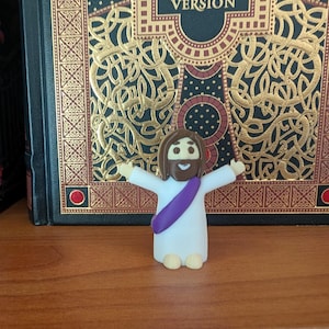 May include: A small, white figurine of a religious figure with outstretched arms, a purple sash, and brown hair and beard. The figurine is in front of a large, ornate book with the words "BIBLE KING JAMES VERSION" on the cover.