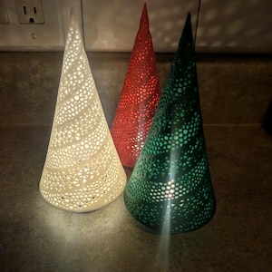 以下が含まれることがあります： クリーム、赤、緑の3つの装飾的な円錐形のキャンドルホルダー。それぞれに光が透過する穴あきデザインが施されています。キャンドルホルダーは暗い表面に配置され、暖かく居心地の良い雰囲気を作り出しています。