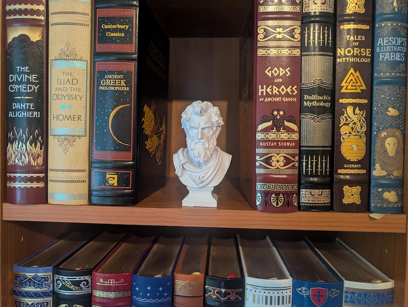 May include: A bookshelf filled with classic literature. The books have decorative spines with titles like "The Divine Comedy," "The Iliad and the Odyssey," and "Gods and Heroes." A white bust of a man sits on the shelf. Below are decorative boxes.