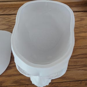 Puede incluir: Un recipiente blanco con forma de calavera, con un dise&ntilde;o en espiral en la base. El recipiente est&aacute; hecho de un material liso y se encuentra sobre una superficie de madera. La calavera tiene detalles faciales.