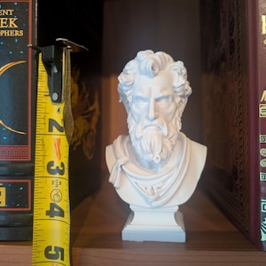 May include: A white bust of a bearded man with curly hair and a draped garment. The bust is approximately 4 inches tall and sits on a square base. It is displayed between two books with gold lettering. One book is titled "Ancient Greek Philosophers".