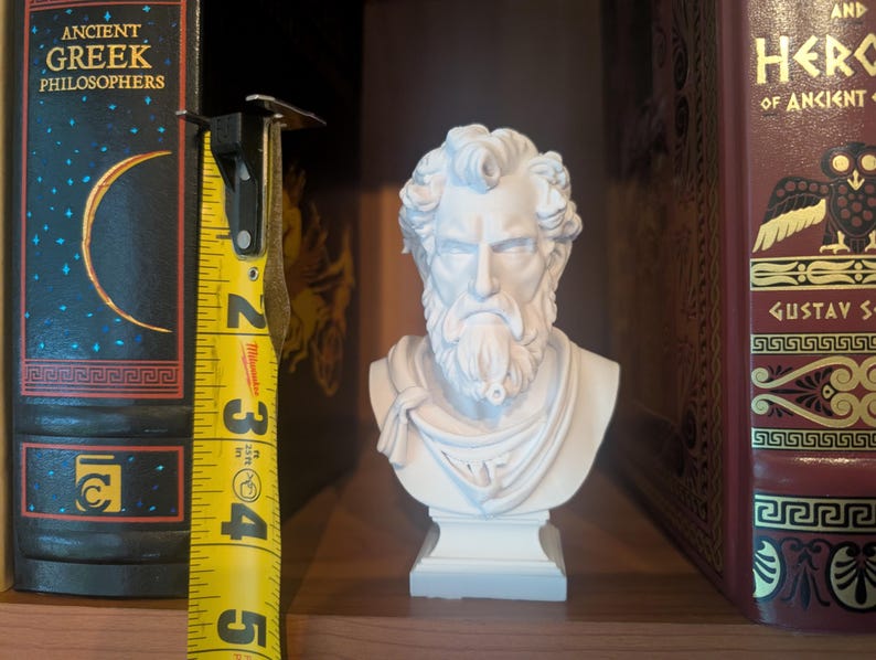 May include: A white bust of a bearded man with curly hair and a draped garment. The bust is approximately 4 inches tall and sits on a square base. It is displayed between two books with gold lettering. One book is titled "Ancient Greek Philosophers".