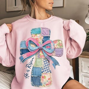 Puede incluir: Una sudadera rosa claro con un diseño de cruz de retazos y un lazo grande. La cruz está hecha de varios patrones de tela, incluyendo floral y mezclilla. Las palabras "He is Risen" están escritas en la sudadera.