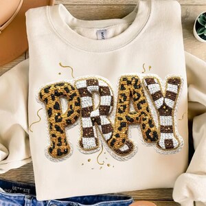 Puede incluir: Sudadera color crema con la palabra "PRAY" en una fuente decorativa. Las letras combinan estampado de leopardo y un patrón a cuadros marrón y blanco, con detalles dorados.
