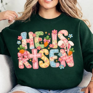 Puede incluir: Sudadera verde oscuro con la frase "HE IS RISEN" en letras de patchwork. Las letras están decoradas con flores, zanahorias y orejas de conejo, ideal para las celebraciones de Pascua.