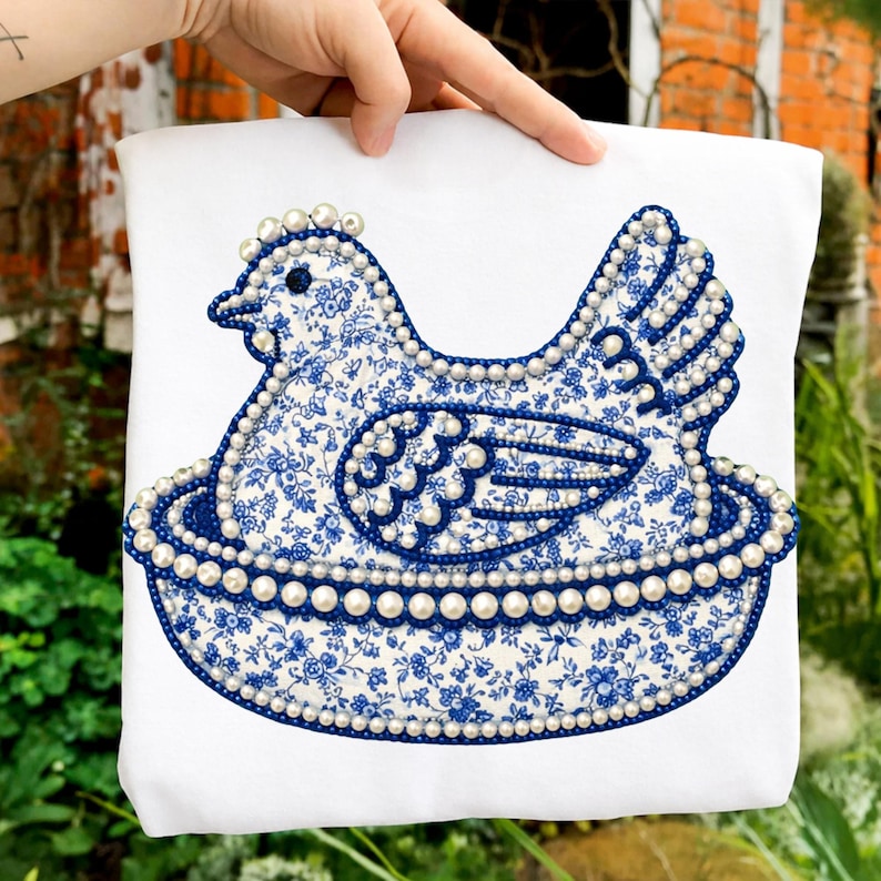 May include: White fabric with a blue floral design hen and nest applique. The hen and nest are outlined with faux pearls. The hen's body is a blue floral pattern. The nest is a blue scalloped design.