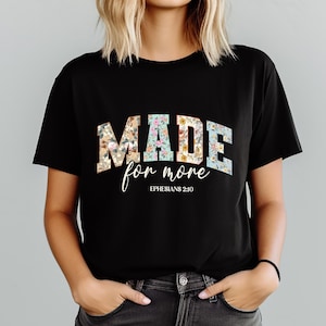 May include: Black t-shirt with the words "MADE for more" in a floral design. The text "Ephesians 2:10" is below the main text in a script font. The shirt is a crew neck style.