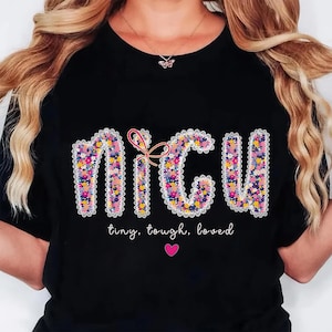 May include: Black t-shirt with the letters "NICU" in a floral design, outlined with white lace. A pink bow is between the "N" and "C". Below the letters, the words "tiny, tough, loved" are written, with a pink heart below.