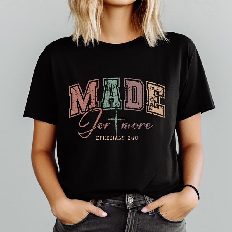 May include: Black t-shirt with the words "MADE for more" in a distressed, colorful font. Below the text is a cross and the words "Ephesians 2:10". The shirt is a crew neck and has short sleeves.