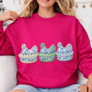 May include: A fuchsia sweatshirt featuring three embroidered hen designs. Each hen is perched on a decorative basket with pearl-like accents. The hens have blue floral patterns and are in different colors.