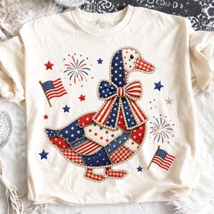 Puede incluir: Una camiseta color crema con el gráfico de un pato decorado con un patrón de patchwork patriótico de estrellas, rayas y cuadros rojos, blancos y azules. El pato lleva un lazo grande y está rodeado de pequeñas banderas y fuegos artificiales.