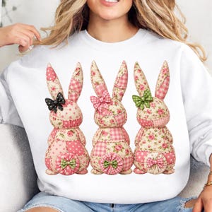 Puede incluir: Sudadera blanca con tres figuras decorativas de conejos. Cada conejo tiene patrones de patchwork en telas rosas, blancas y florales. Los conejos tienen lazos en negro, rosa y verde.
