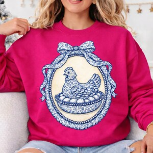May include: A magenta sweatshirt featuring a blue and white floral design of a hen sitting on eggs within an oval frame. The frame is adorned with a bow and ribbon, and the design is reminiscent of vintage porcelain.