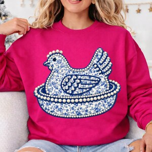 May include: A fuchsia sweatshirt featuring a blue and white floral patterned hen design. The hen is sitting in a nest, and the design is outlined with faux pearls. The sweatshirt has long sleeves.