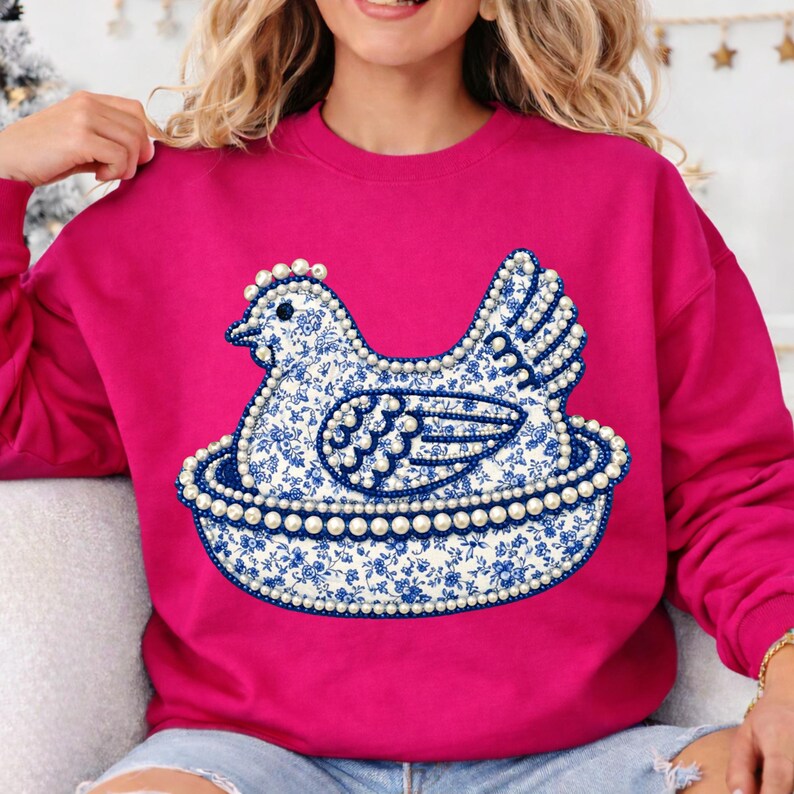 May include: A fuchsia sweatshirt featuring a blue and white floral patterned hen design. The hen is sitting in a nest, and the design is outlined with faux pearls. The sweatshirt has long sleeves.