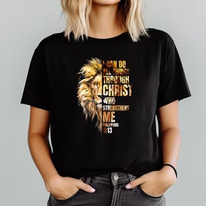 Może przedstawiać: Czarny t-shirt z złotym wzorem głowy lwa po lewej stronie. Tekst "I CAN DO ALL THINGS THROUGH CHRIST WHO STRENGTHENS ME PHILIPPIANS 4:13" jest wydrukowany w kolorze złotym i białym.